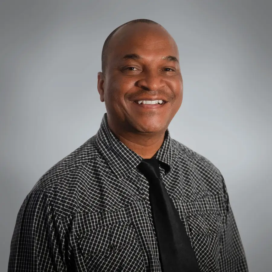 Smiling man in a black-and-white plaid shirt and black tie, posed against a gray studio backdrop.