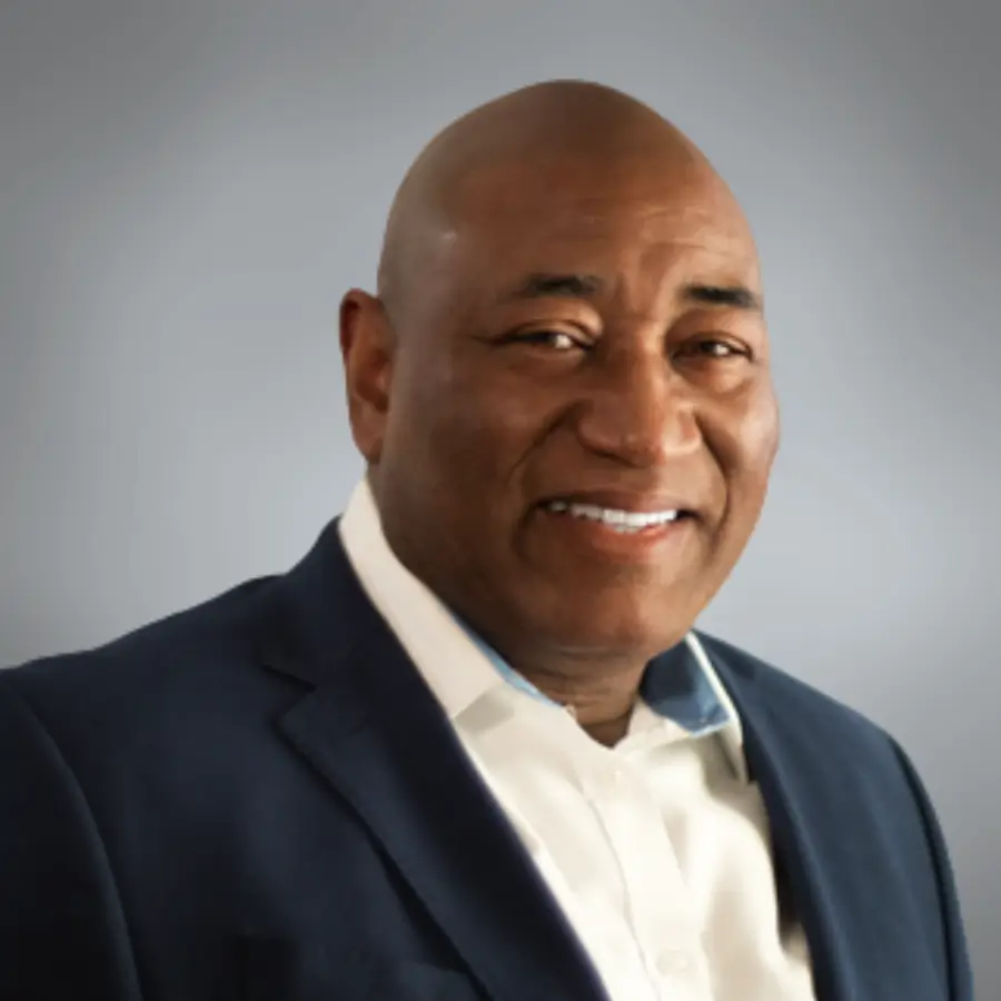 Smiling bald man in a dark blazer and white shirt poses for a professional headshot against a gray backdrop.