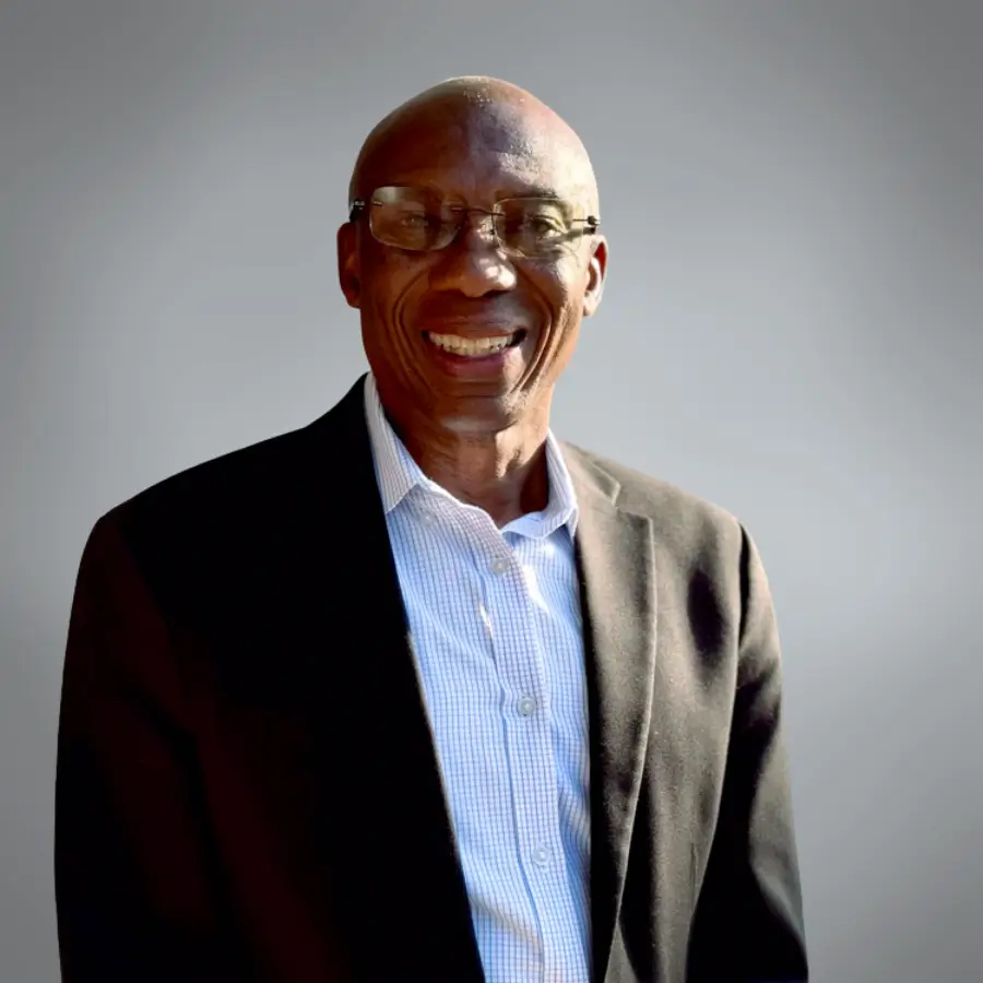 Smiling man in a dark blazer and light blue checkered shirt, wearing glasses, posed against a neutral gray background.