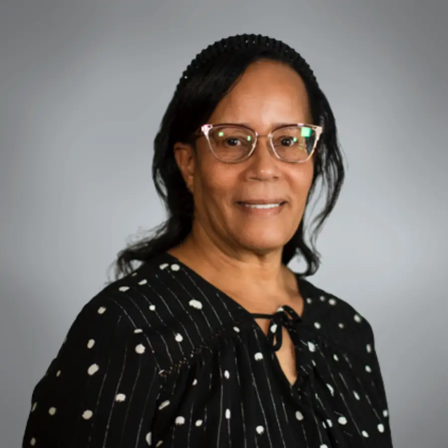 Portrait of a middle-aged woman with glasses, wearing a black polka-dot blouse, smiling against a neutral gray background.